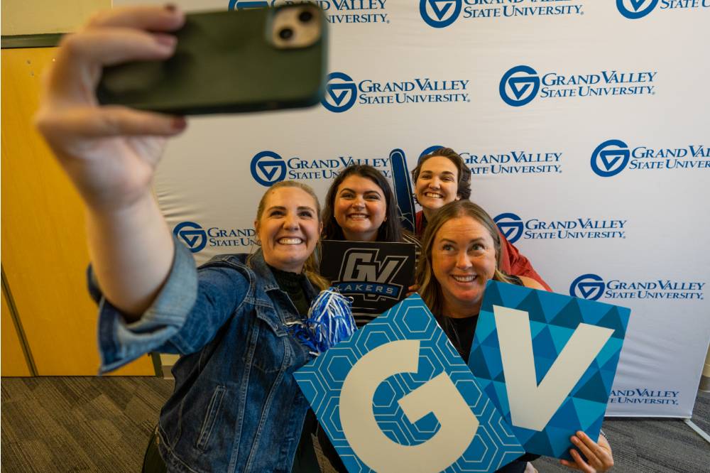 Group photo: someone taking a selfie in front of GVSU banner, the group posing with foam fingers and the GV signs
